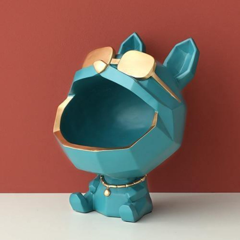 A geometric dog-shaped decorative planter in teal blue with gold trim and gold-toned sunglasses, shown on a white surface against a muted red-brown background.