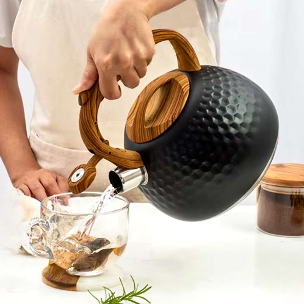 Person pouring tea from a black teapot with wooden handle into a glass mug on a white background.
