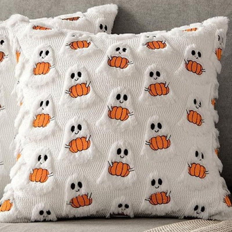 A white textured throw pillow with a fuzzy fringe edge, featuring a repeating pattern of embroidered orange pumpkins with black eyes and mouths surrounded by small white ghosts.