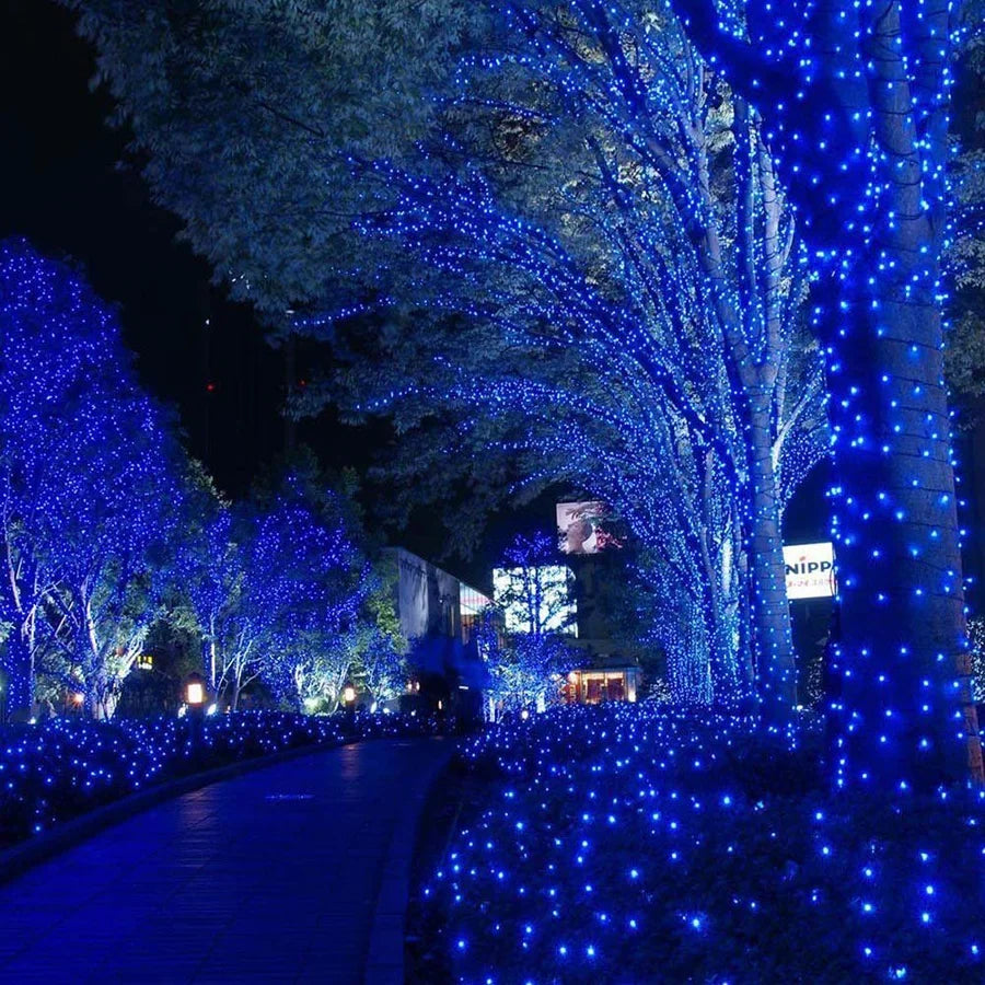 Blue LED string lights wrapped around trees and bushes along a nighttime pathway, creating a glowing blue canopy in an outdoor park setting.