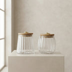 Two ribbed clear glass jars with light wood lids and small gold metal pulls, displayed on a beige pedestal against a neutral beige wall.