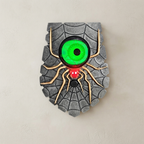 A Halloween-style wall decoration shaped like a spider on a web, mounted on a textured beige wall. The design features a gray webbed shield background, tan spider legs, a bright green glowing eye, and a red light at the bottom.
