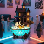 A Halloween-themed decorative scene with a small ghost character wearing a witch hat sitting in a glowing blue mosaic bath bowl and reading a book, set in front of a miniature haunted house with skeleton props and tombstones.