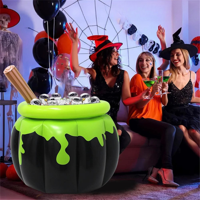 A large inflatable cauldron-shaped party table with bright green slime-like drips and silver cup holders, shown with three women in Halloween costumes sitting on a couch in a decorated living room.