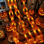 A set of glowing orange LED chain lights hanging in the foreground, with carved jack-o'-lantern pumpkins and Halloween decorations arranged on a table in a decorated indoor setting.