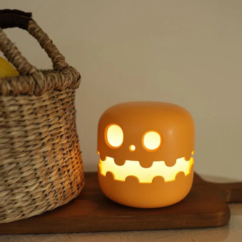 A glowing orange jack-o'-lantern-shaped night light with a cute cartoon face, sitting on a wooden board next to a woven basket.