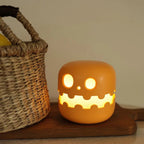 A glowing orange jack-o'-lantern-shaped night light with a cute cartoon face, sitting on a wooden board next to a woven basket.