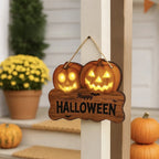 A wooden Halloween hanging sign with two glowing jack-o'-lantern faces and the words 'Happy HALLOWEEN' in black, suspended by a rope in front of a porch door. Pumpkins and potted flowers are visible in the background.