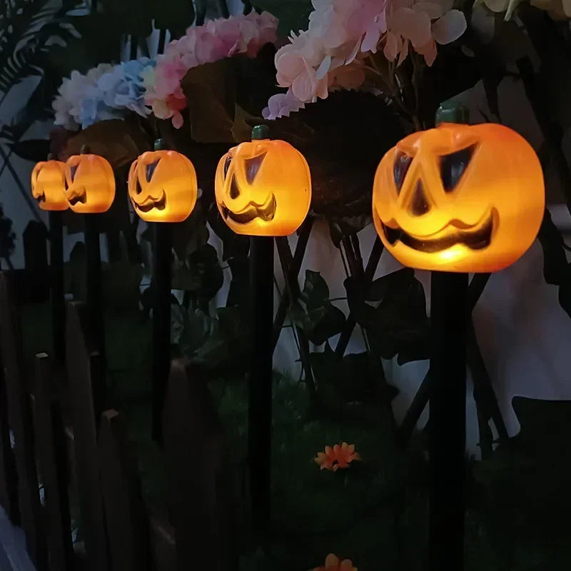 Five glowing jack-o'-lantern Halloween yard lights mounted on black stakes in a garden setting with artificial flowers and a dark fence in the background.