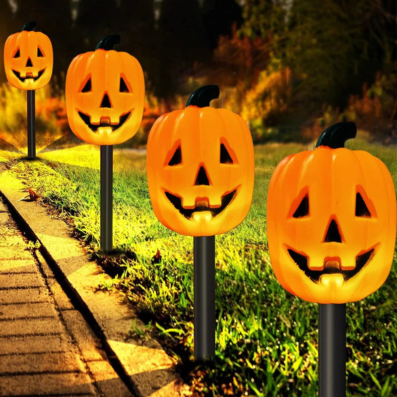 Four glowing jack-o'-lantern Halloween yard lights on black stake posts, arranged along a grassy lawn next to a paved walkway at night.