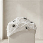 White faux spiderweb draped over a box, decorated with several black plastic spiders, set against a neutral beige background.