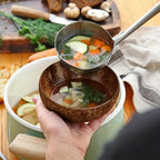 Hand holding a coconut bowl with vegetable soup, ladle pouring into it, on a wooden surface.