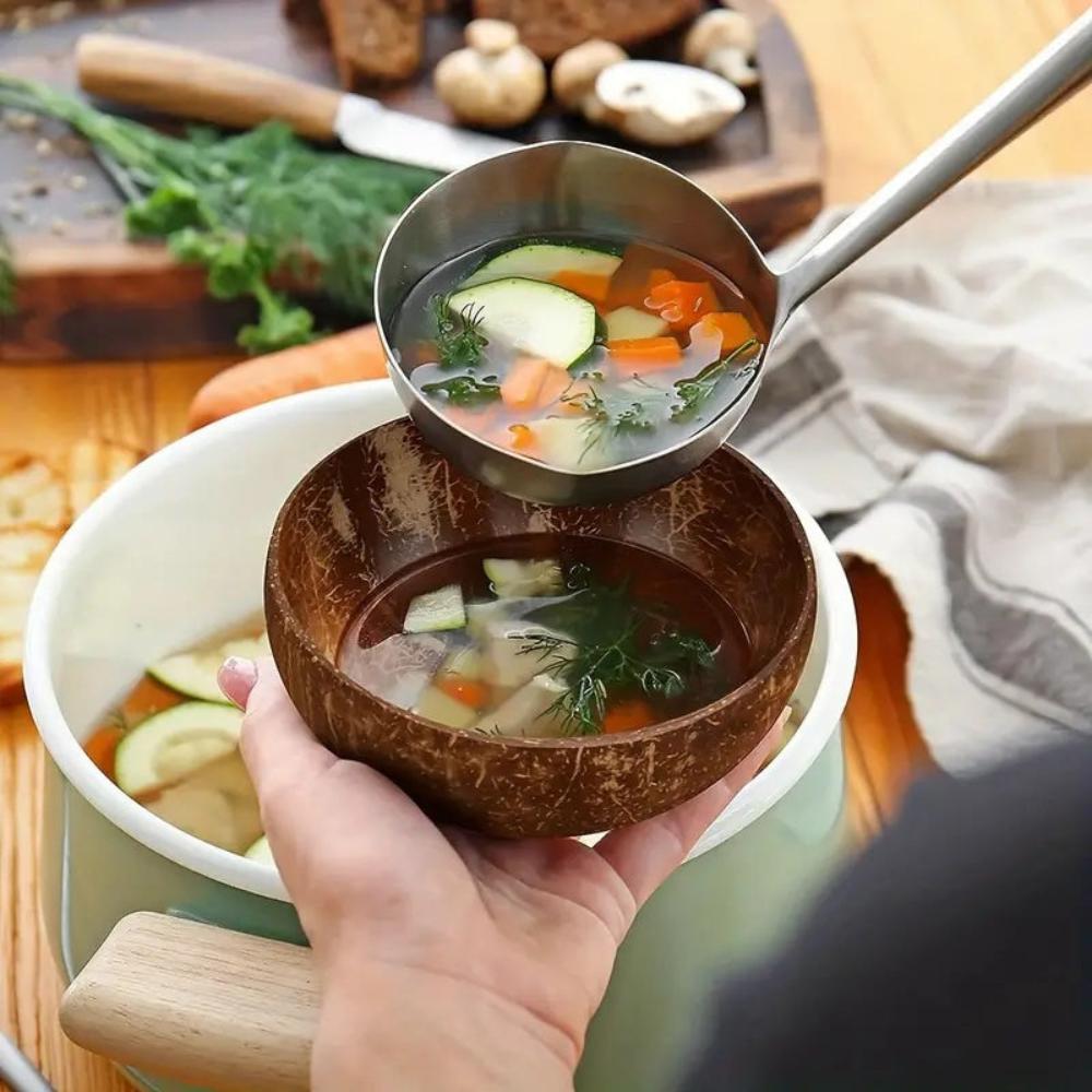 Hand holding a coconut bowl with vegetable soup, ladle pouring into it, on a wooden surface.