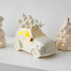 White ceramic car with a Christmas tree on top, surrounded by other decorative items on a light gray background.