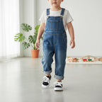 A toddler walking indoors wearing blue denim overall-style pants over a white T-shirt, with black-and-white sneakers. The room has a light gray floor, white walls, a potted plant, and a small wooden toy setup in the background.