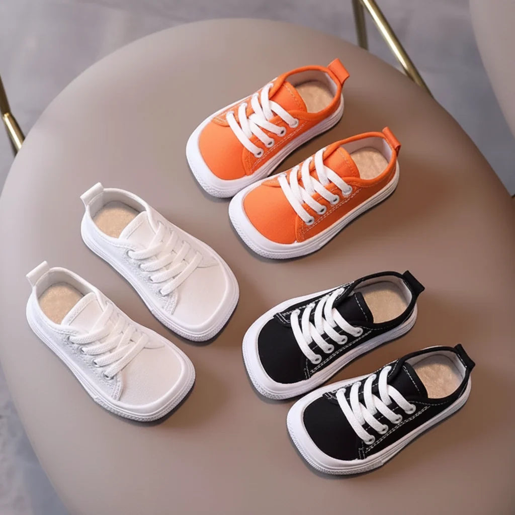 Four low-top canvas lace-up shoes arranged on a round taupe stool: two orange shoes, two black shoes, and one white shoe from each colorway. The shoes have white laces and white rubber soles.