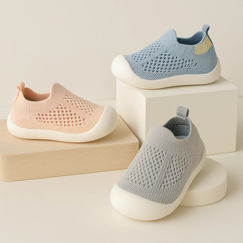 Three knit slip-on baby shoes in pastel pink, light blue, and light gray, displayed on minimalist beige blocks against a warm off-white background.