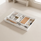 A white rectangular kitchen utensil organizer tray with multiple compartments, shown holding cutlery, chopsticks, and small kitchen tools on a light beige tabletop in a softly lit room.