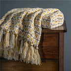 Yellow and gray patterned blanket with tassels draped over a wooden bench against a gray wall.