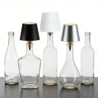 Clear glass bottles with black, white, and silver lampshades on a reflective surface.