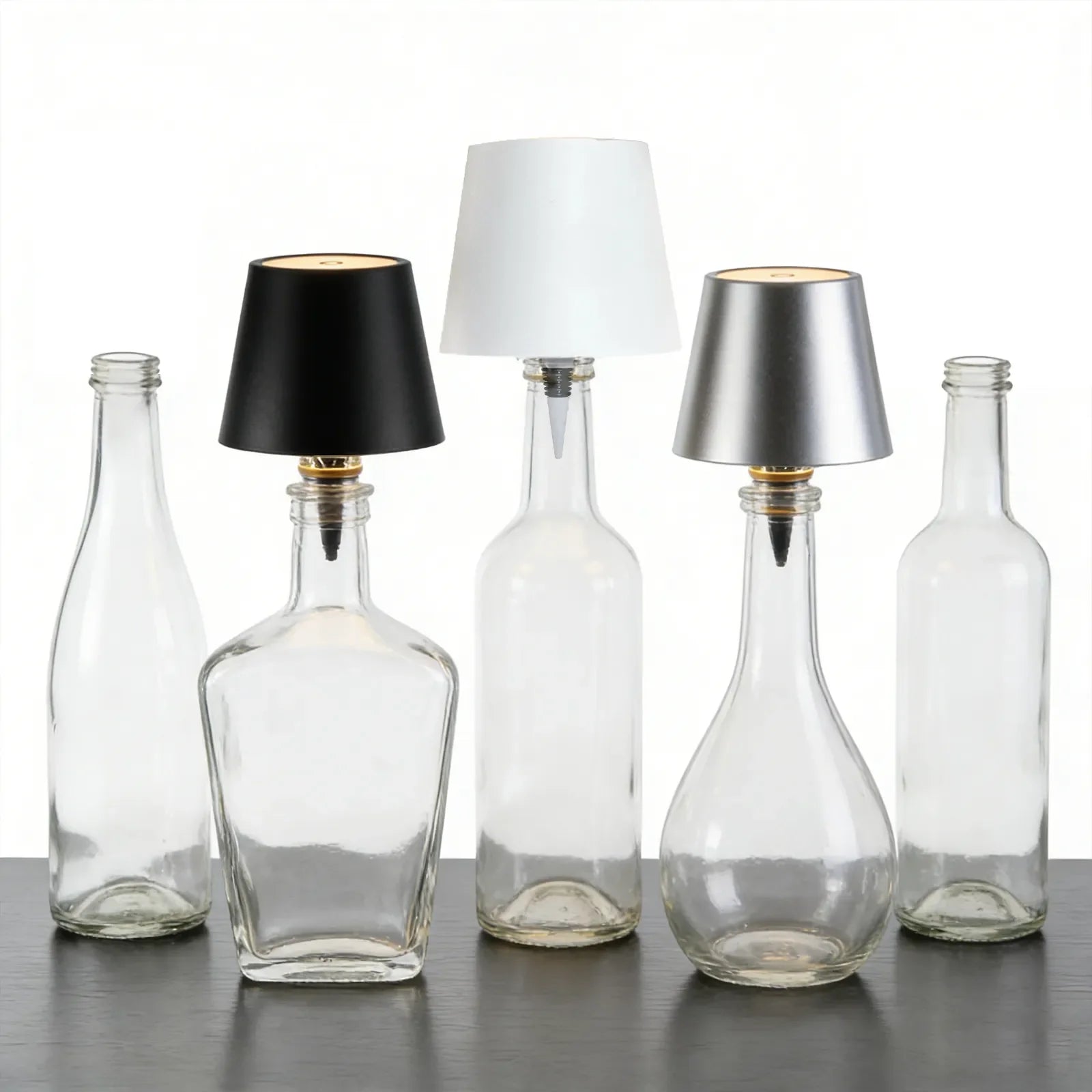 Clear glass bottles with black, white, and silver lampshades on a reflective surface.