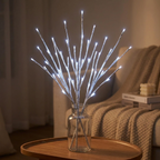 White LED branch-style string lights in a clear glass vase, displayed on a wooden table in a cozy living room setting.