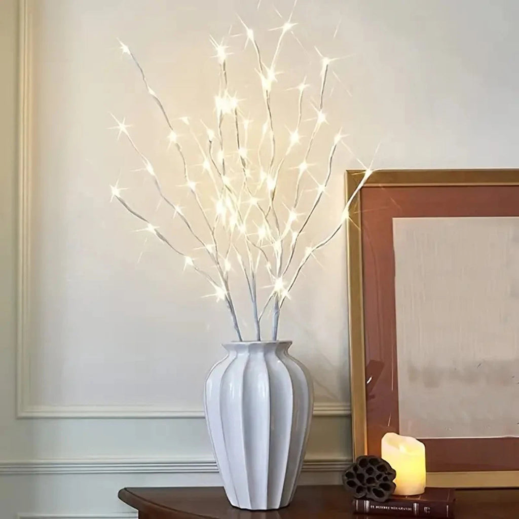 White vase with illuminated branches on a table against a wall with a framed picture.