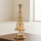 A small gold wire spiral Christmas tree with warm white string lights and a gold star topper, displayed on a wooden surface in a softly lit indoor setting.
