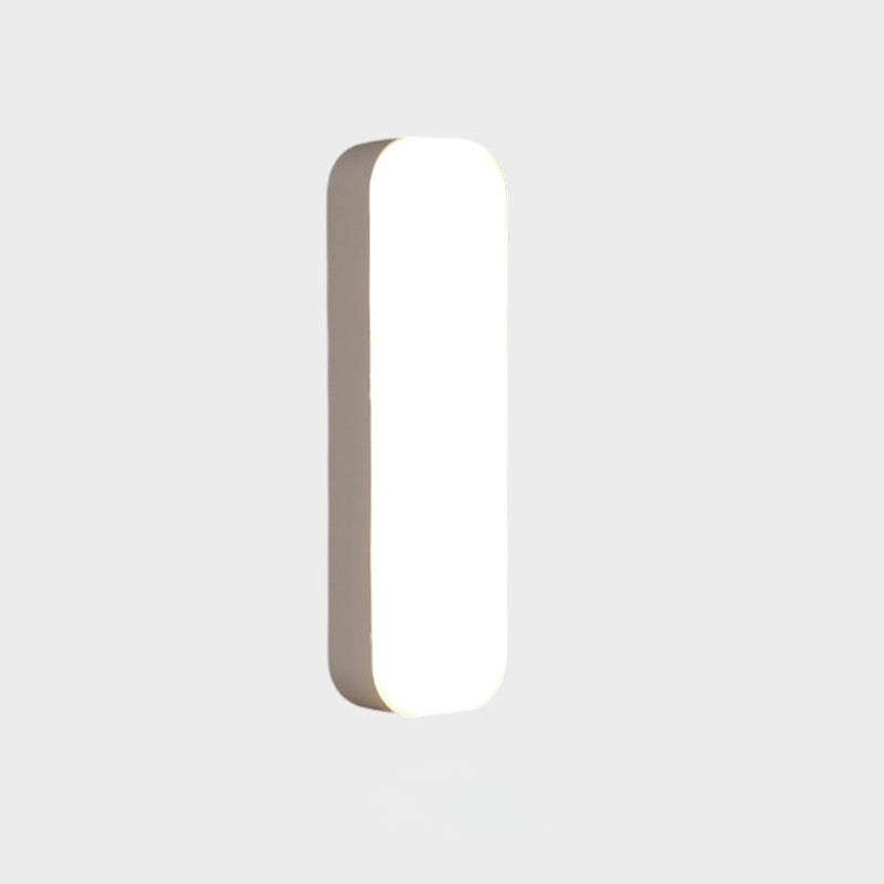 A modern, minimalist vertical rectangular wall light with rounded corners, shown illuminated against a solid light gray background.