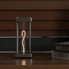 A modern table lamp with a square dark metal frame and a glowing coiled Edison-style bulb inside, placed on a wooden surface next to stacked books against a dark wood slat wall.