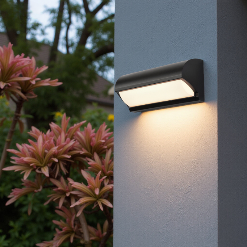 Black outdoor wall light fixture on a gray wall with pink flowers in the foreground.