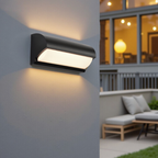 Modern outdoor wall light fixture on a gray wall with a patio and seating area in the background.