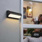 Black outdoor wall light fixture on a gray wall with a blurred indoor scene in the background.