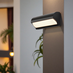 Modern outdoor wall light fixture on a building exterior with plants in the background.