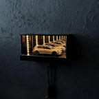 A rectangular black wall-mounted display box with a glowing interior showing a miniature model car in a garage scene, mounted on a dark textured wall.