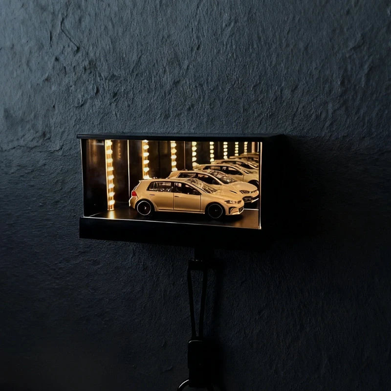 A rectangular black wall-mounted display box with a glowing interior showing a miniature model car in a garage scene, mounted on a dark textured wall.