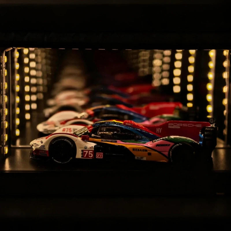A miniature Porsche 911 race car model displayed on a dark shelf in a lit display case, with other race cars blurred in the background.
