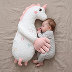 A baby in a light gray ribbed onesie sleeps peacefully while hugging a large white minky dot unicorn plush toy with pink accents, set against a neutral beige fabric background.