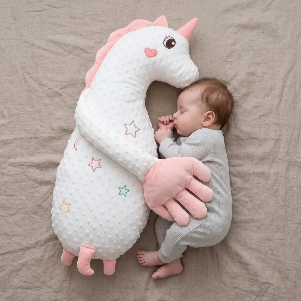 A baby in a light gray ribbed onesie sleeps peacefully while hugging a large white minky dot unicorn plush toy with pink accents, set against a neutral beige fabric background.