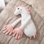A plush unicorn-shaped baby pillow with long arms and pink hooves, resting on a beige linen surface with a cream knit blanket nearby.