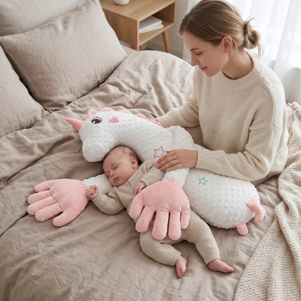 A woman sits on a beige bed beside a sleeping baby, holding a plush unicorn-shaped baby support pillow with pink paw-shaped ends. The scene is softly lit with neutral bedding and a wooden bedside table in the background.