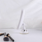 A white adjustable makeup mirror with a suction cup base, shown on a white surface with two makeup brushes in the foreground against a draped white fabric background.