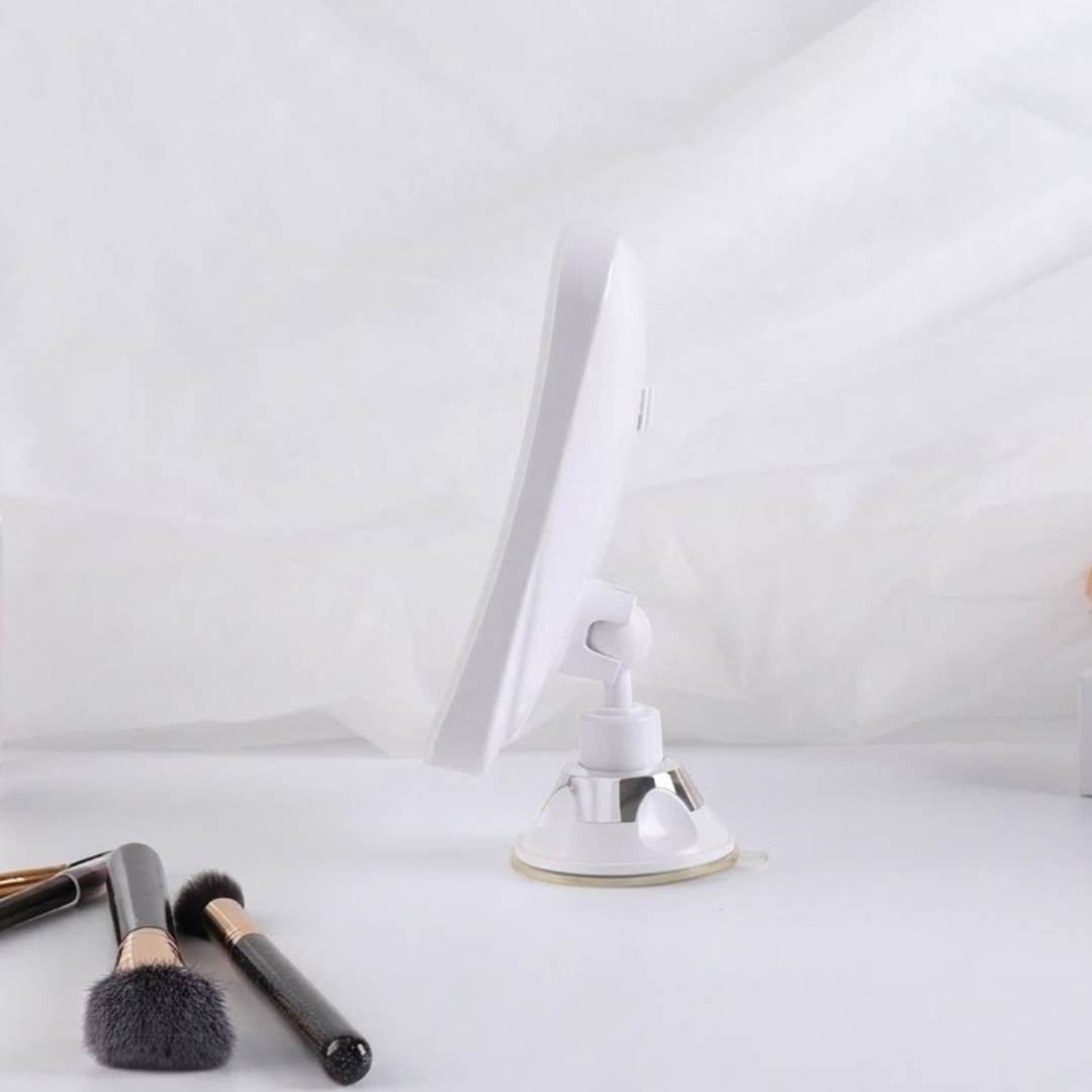 A white adjustable makeup mirror with a suction cup base, shown on a white surface with two makeup brushes in the foreground against a draped white fabric background.