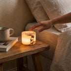 Hand reaching towards a lit candle on a wooden table next to a mug and book, with a cozy indoor setting.