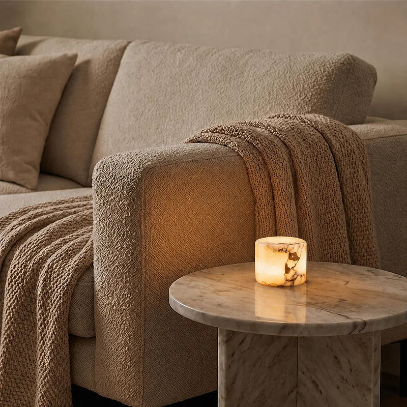 A lit cylindrical candle with a warm glowing amber interior sits on a round marble side table next to a beige textured sofa with a knitted throw blanket.