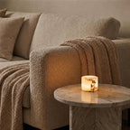 A lit cylindrical candle with a warm glowing amber interior sits on a round marble side table next to a beige textured sofa with a knitted throw blanket.