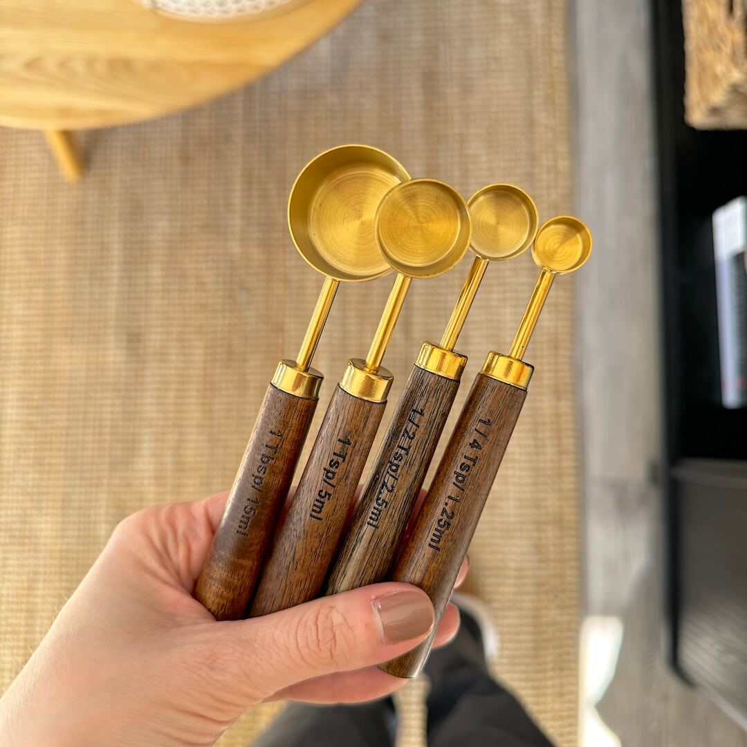 Set of four gold measuring spoons with wooden handles held by a hand against a neutral background.