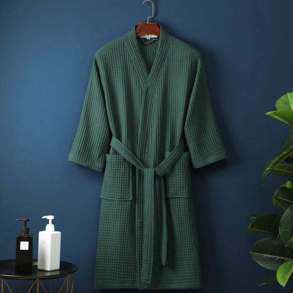 Green bathrobe hanging on a hook against a blue wall with a plant and bottles on a table.