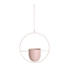 A minimalist hanging planter in a soft dusty pink color, featuring a matte ceramic cup suspended inside a thin circular metal frame with a hanging wire loop and a top suspension cord, set against a solid white background.