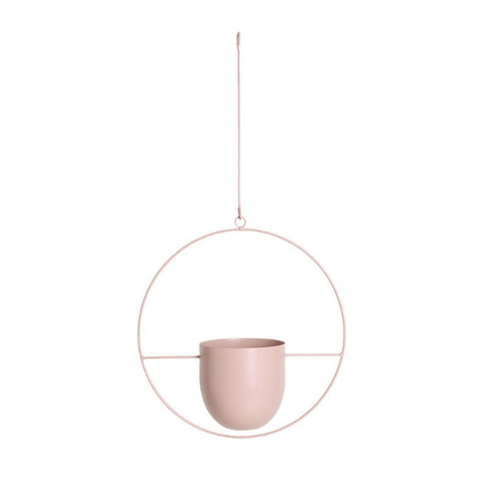 A minimalist hanging planter in a soft dusty pink color, featuring a matte ceramic cup suspended inside a thin circular metal frame with a hanging wire loop and a top suspension cord, shown against a solid white background.